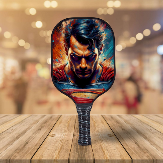 Pickleball paddle with black face featuring Superman splash artwork and blue edge trim